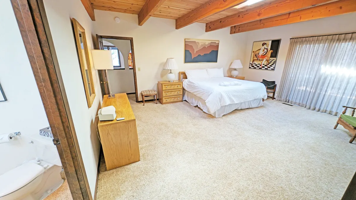 A spacious bedroom anchored by a large king bed, with beautiful artwork on the wall, exposed wooden beams in the ceiling, and soft curtains filtering natural light. The atmosphere is serene and inviting, ideal for restful mornings or cozy evenings.