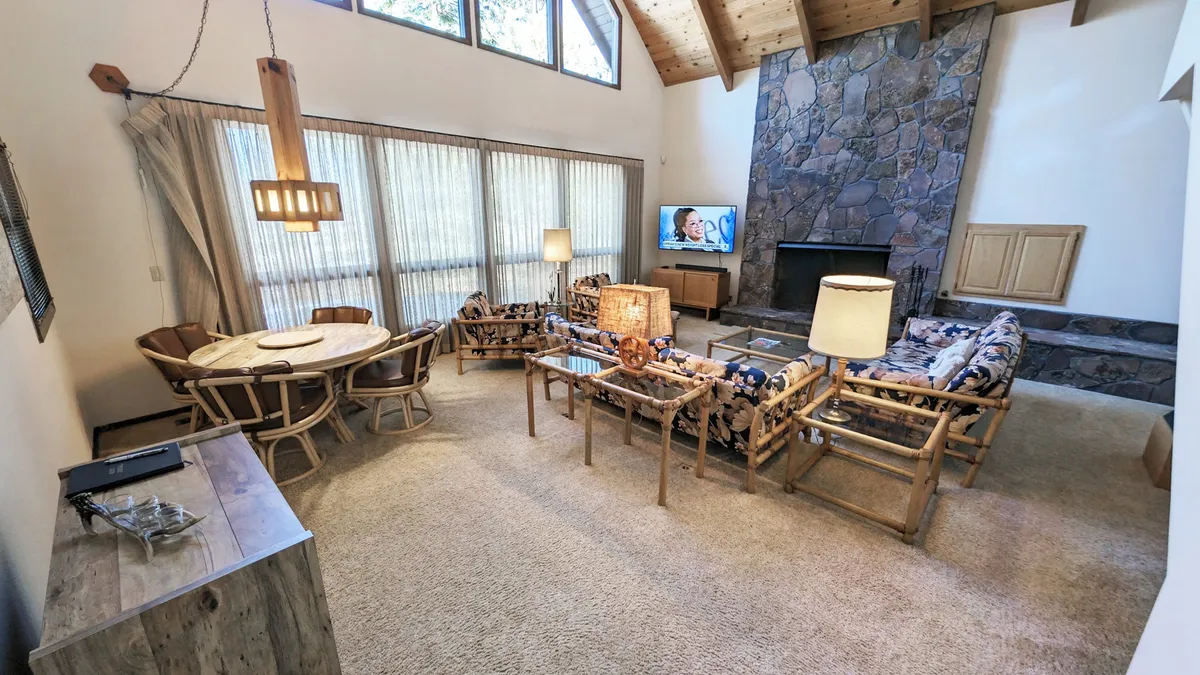 The image showcases a spacious and sunlit living room with a striking stone fireplace, expansive windows that flood the space with natural light, comfortable wooden furniture, and a large TV set. The layout suggests an ideal setting for family gatherings, cozy evenings, or leisurely mornings, emphasizing warmth and togetherness.