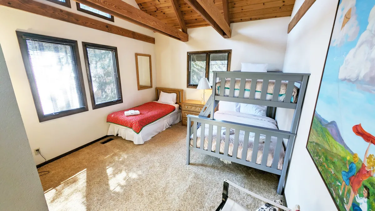 A lively and colorful bedroom featuring a bunk bed and a single bed, bright wall art, and large windows letting in abundant morning sunlight. The space feels playful, perfect for sleepovers, games, and storytelling.