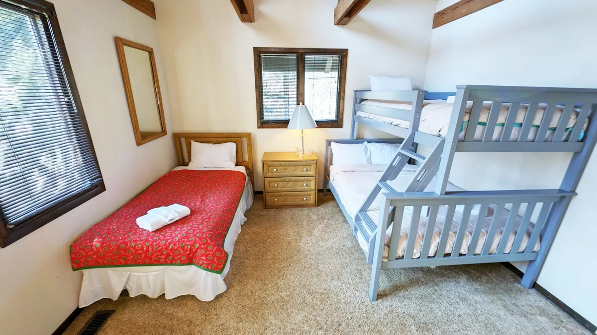 A warm and inviting bedroom space with a bunk bed, single bed, lamp, and a nightstand by the window. The natural light highlights the cozy arrangement, creating the perfect spot for late-night storytelling or relaxed family moments.