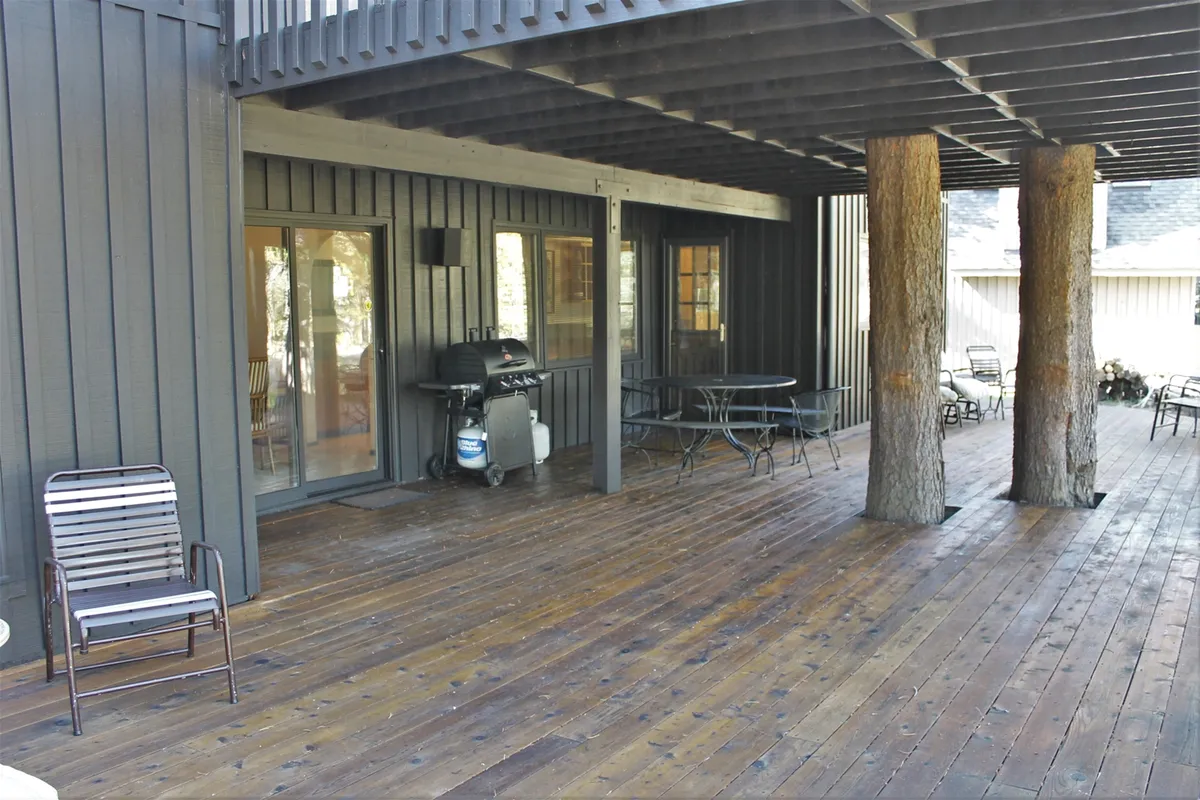 This image captures a covered outdoor deck with wooden pillars, a comfortable seating area, a barbecue grill, and a large table. The setting is partially sheltered, surrounded by lush trees, and offers ample space for gatherings and relaxation.
