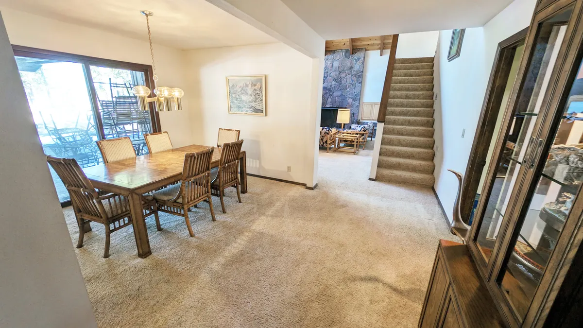 A warm and inviting dining area centered around a large table, situated near a stone fireplace and adjacent to a visible staircase and sliding glass doors. The space is designed for group meals, celebrations, and relaxing together, with light streaming in from the doors.