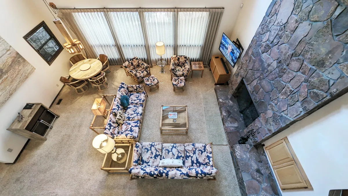 This image features a spacious living-dining area with a large stone fireplace, floral patterned sofas, a circular dining table, and a television. The room's layout is open and communal, perfect for gathering the whole family—including pets—after spending time exploring outside.