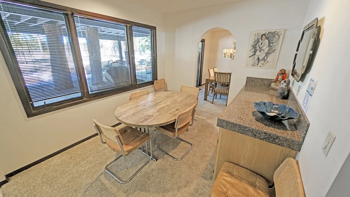 Inside, a sunlit dining area features a round wooden table surrounded by stylish chairs, large windows with blinds that bring in natural light, a granite countertop, and a decorative bowl. The space feels inviting and ideal for family meals or casual gatherings after a day of activities.