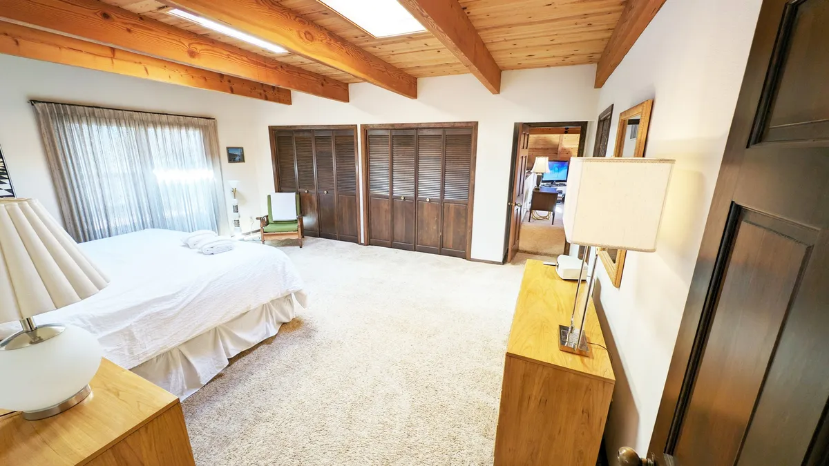 A cozy bedroom featuring a bed with white bedding, wooden furniture, and large windows with sheer curtains that let in morning sunlight. Outside the windows, greenery hints at a natural setting.
